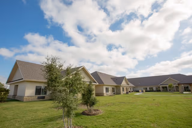 Exterior view of a single-story healthcare and rehabilitation facility building with beige siding and stone accents, surrounded by a well-maintained green lawn and a few small trees under a partly cloudy sky.