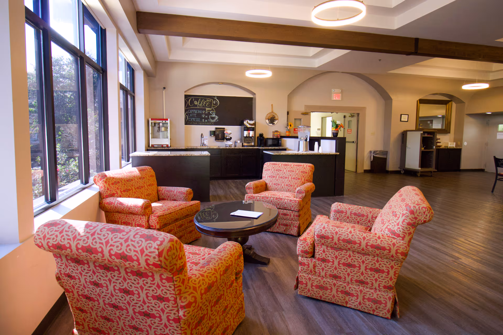 A cozy seating area with four patterned armchairs arranged around a round wooden table in a well-lit room with large windows. In the background, there is a coffee station with a chalkboard menu, coffee machines, and a popcorn machine. The room has wooden flooring and modern circular ceiling lights.