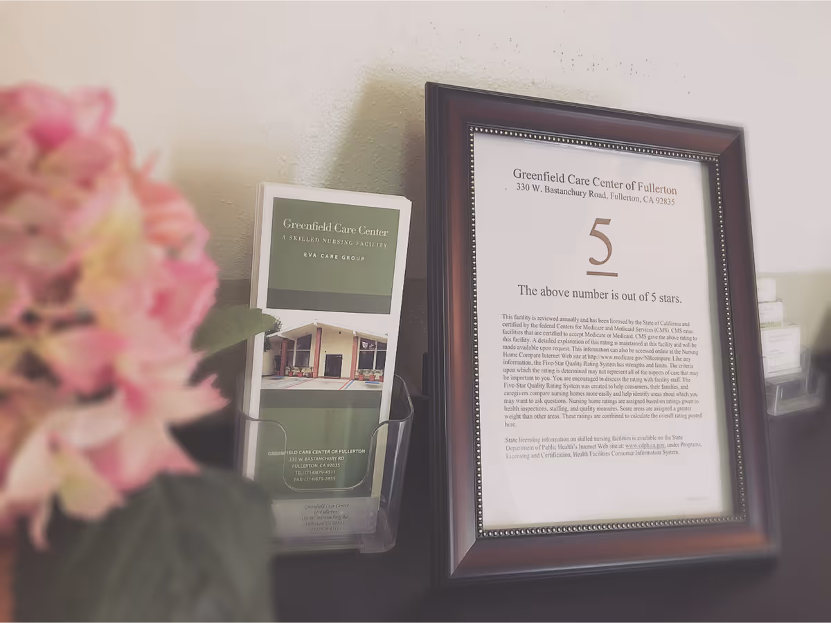 A framed certificate from Greenfield Care Center of Fullerton displaying a 5-star rating, placed on a dark surface next to a brochure holder containing brochures for the facility. A pink flower is partially visible in the foreground on the left side.