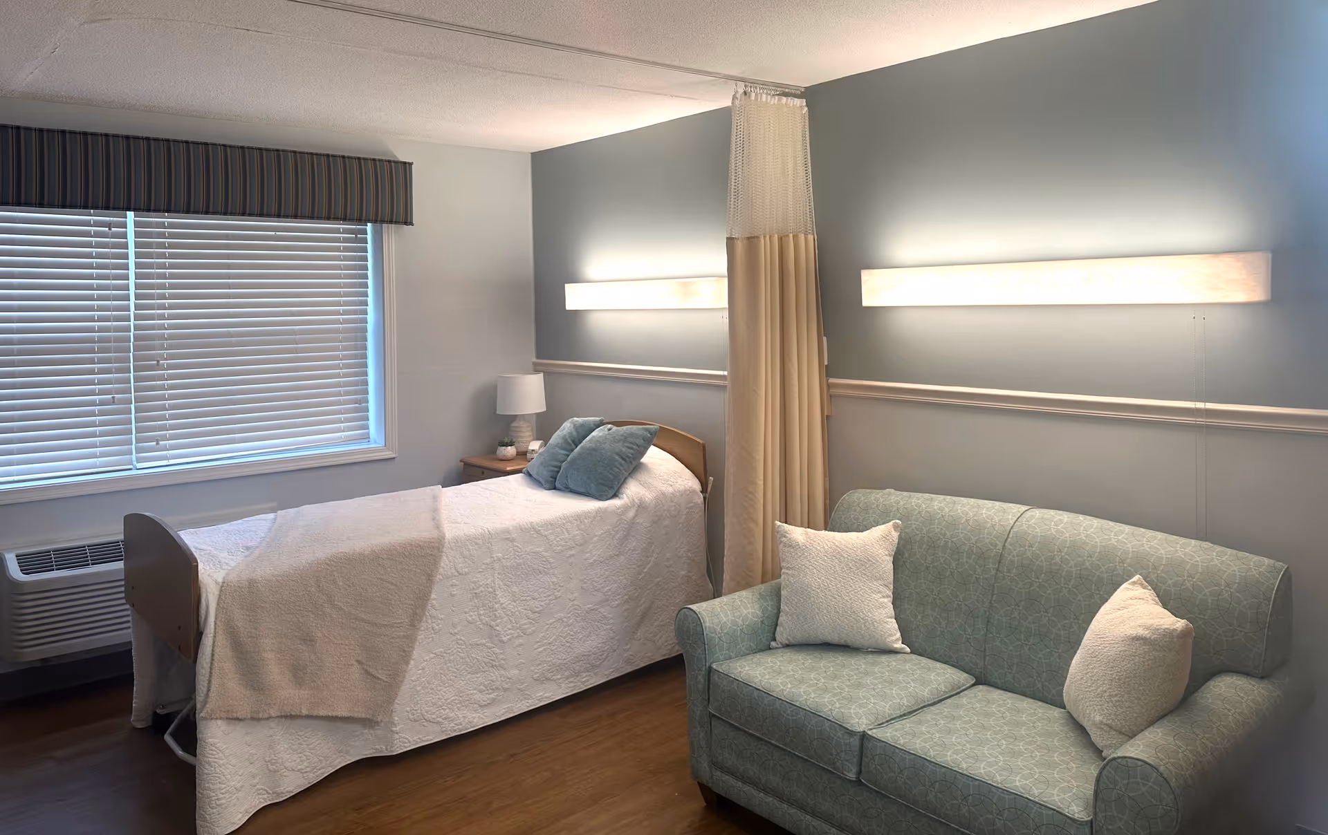 A cozy room in a rehabilitation center featuring a single bed with white bedding and two blue pillows, a small wooden nightstand with a lamp, a green patterned couch with two white pillows, a window with closed blinds and a striped valance, and a privacy curtain hanging from the ceiling. The walls are painted light blue and white, and there are two horizontal wall lights above the bed and couch.