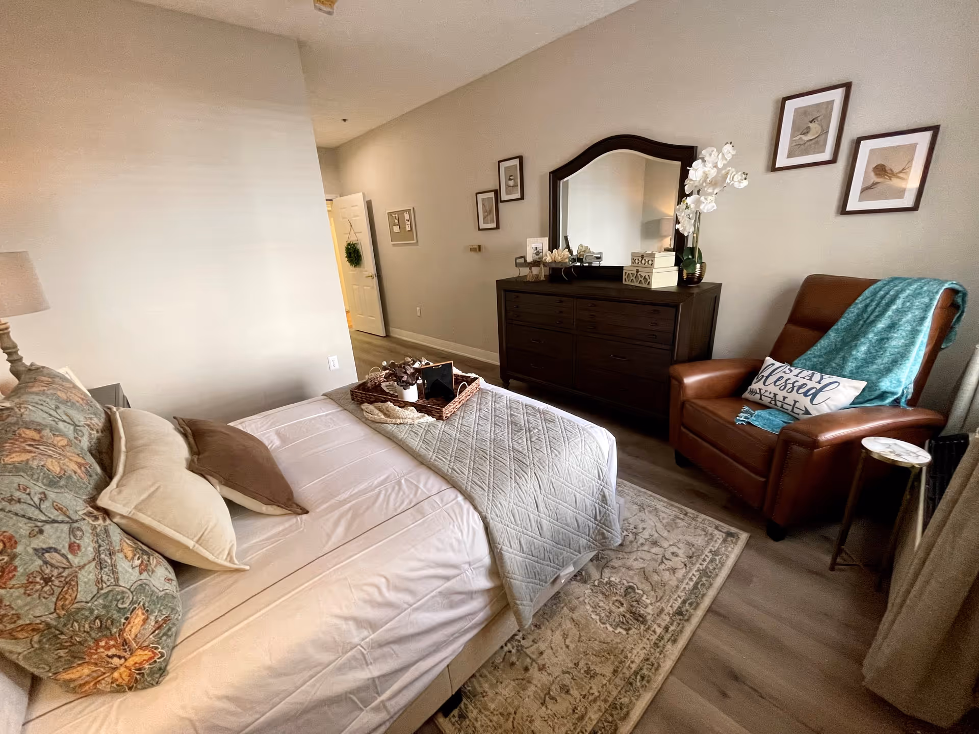 A cozy bedroom featuring a neatly made bed with multiple pillows and a quilted blanket. A dark wooden dresser with a large mirror sits against the wall, decorated with a white orchid plant and small decorative items. To the right, there is a brown leather armchair with a teal throw blanket and a pillow that reads 'Stay Blessed Y'all'. The room has light-colored walls, framed bird artwork, and a patterned area rug on the wooden floor.