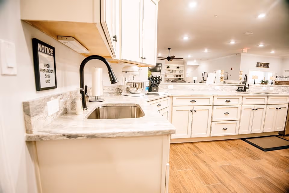 A bright and clean kitchen area with white cabinets, marble countertops, and a stainless steel sink with a black faucet. There is a stand mixer, a knife block, and a paper towel holder on the counter. A sign on the wall reads 'NOTICE FOOD PREPARATION SINK ONLY'. The floor is wooden, and the background shows a spacious, well-lit room with ceiling lights and a ceiling fan.