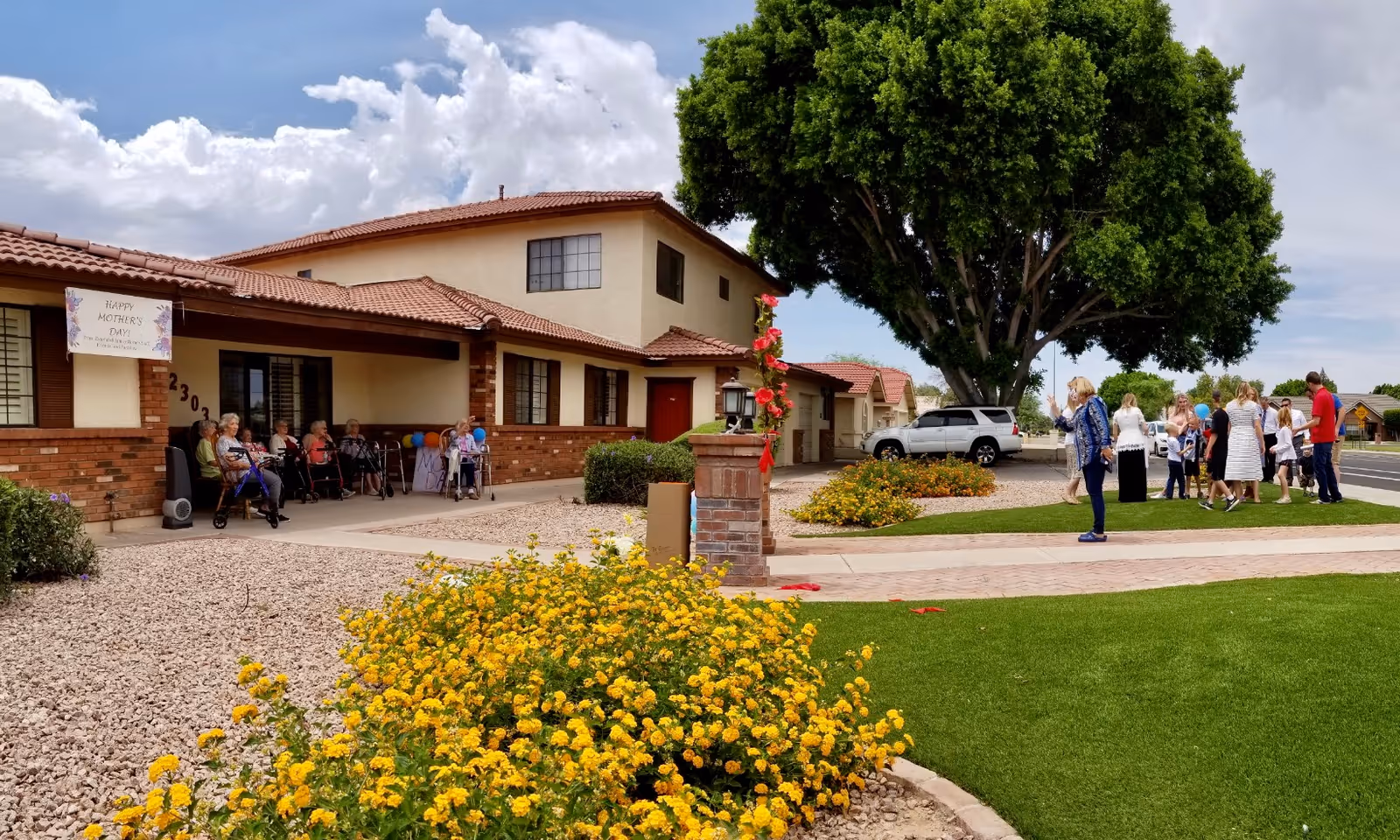 Outdoor scene at Extended Family Home - West showing a group of elderly residents sitting on chairs under a covered porch with a Happy Mother's Day banner. In the foreground, there are yellow flowers and a well-maintained lawn. To the right, a group of people, including children and adults, are gathered near a large tree and parked cars.