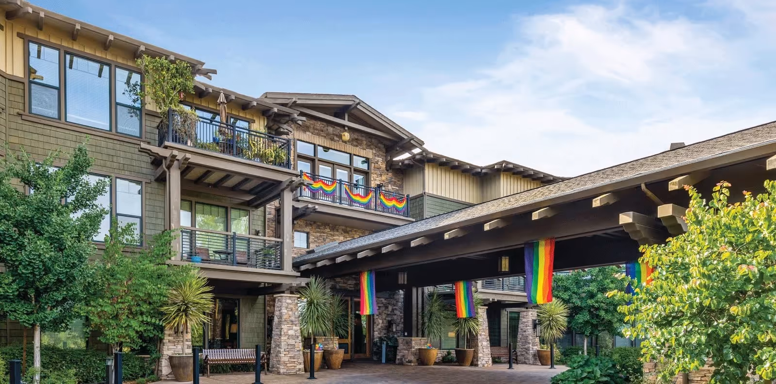 Exterior view of a senior living facility building with multiple balconies decorated with rainbow flags. The building has stone and wood siding, surrounded by green trees and plants under a partly cloudy sky.