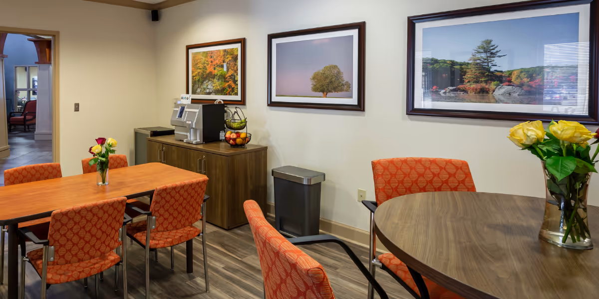 Interior view of a dining area in The Lodge at Natchez Trace featuring two wooden tables with orange patterned chairs. One table has a vase with yellow and red roses, and the other table has a vase with yellow roses. A wooden cabinet against the wall holds a coffee machine and a fruit basket. Three framed landscape photographs hang on the wall above the cabinet. The floor is wood-patterned, and there is a trash bin next to the cabinet.