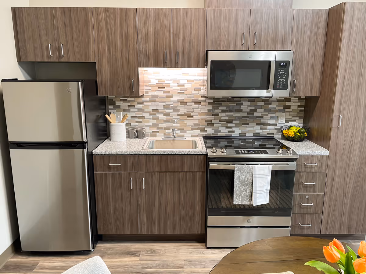 Modern kitchen area with stainless steel refrigerator, microwave, and oven. The kitchen features wood grain cabinets, a tiled backsplash in neutral tones, a small sink, and a countertop with kitchen utensils and a bowl of fruit. Part of a round wooden table with orange flowers is visible in the foreground.