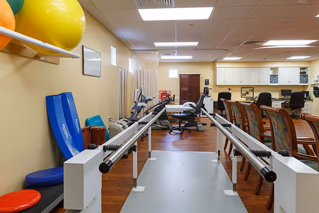 A rehabilitation or physical therapy room with parallel bars for walking exercises in the center. The room has exercise equipment including stability balls, exercise mats, and stationary bikes. There is a row of chairs and a desk area with computers and office chairs in the background. The floor is wooden, and the walls are painted light yellow.