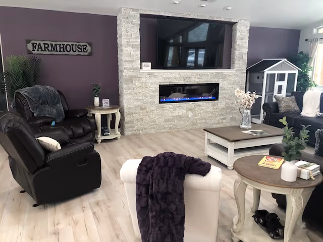 A cozy living room with light wood flooring, a stone fireplace with a mounted flat-screen TV above it, and comfortable seating including a black recliner, a white armchair with a dark purple throw, and a dark sofa. There are side tables with decorative items, a coffee table, a small indoor playhouse, and a wall sign that reads 'FARMHOUSE'.