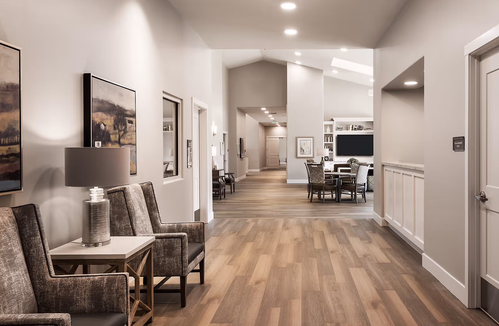 A spacious, well-lit hallway in an assisted living facility with wood flooring, neutral-colored walls, and modern decor. On the left side, there are two upholstered chairs with a side table and a lamp between them, along with framed artwork on the wall. Further down the hallway, there is a seating area with a round table and chairs, a large flat-screen TV mounted on the wall, and built-in shelves with decorative items.