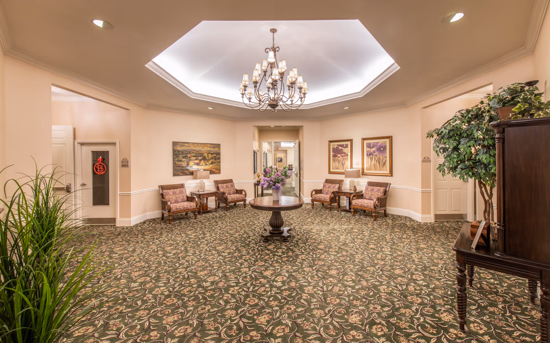 A furnished senior living lobby with patterned carpet, a central table with flowers, seating areas, framed art, and a chandelier.