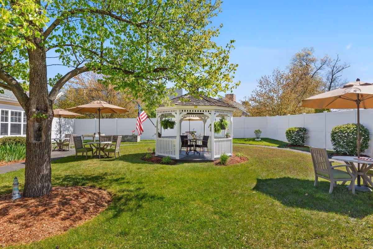 A sunny outdoor garden area with green grass, a large tree, and a white gazebo in the center. Several tables with chairs and umbrellas are placed around the garden. An American flag is visible near the gazebo, and a white fence encloses the area.