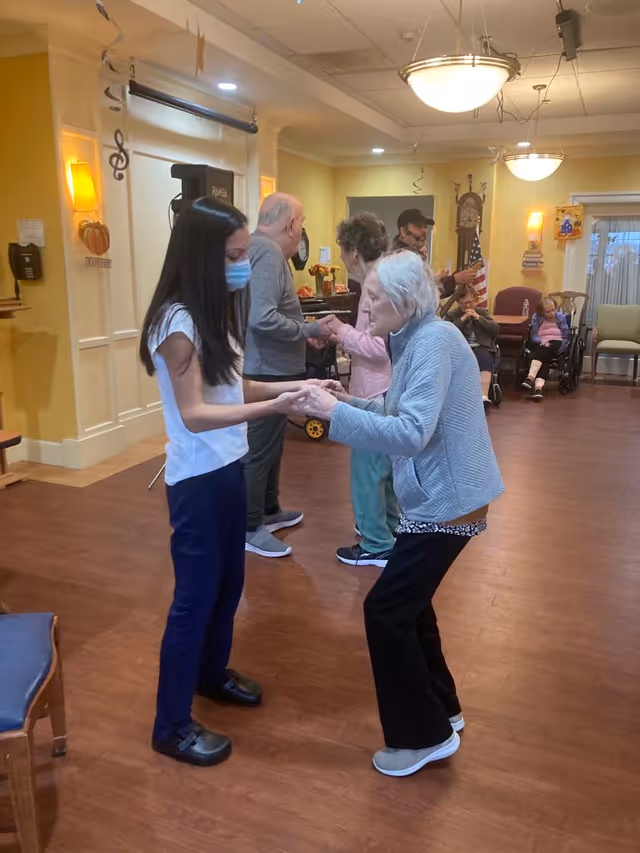 A masked staff member holds hands and dances with an elderly resident in a senior living facility common room while other residents sit and socialize in the background.