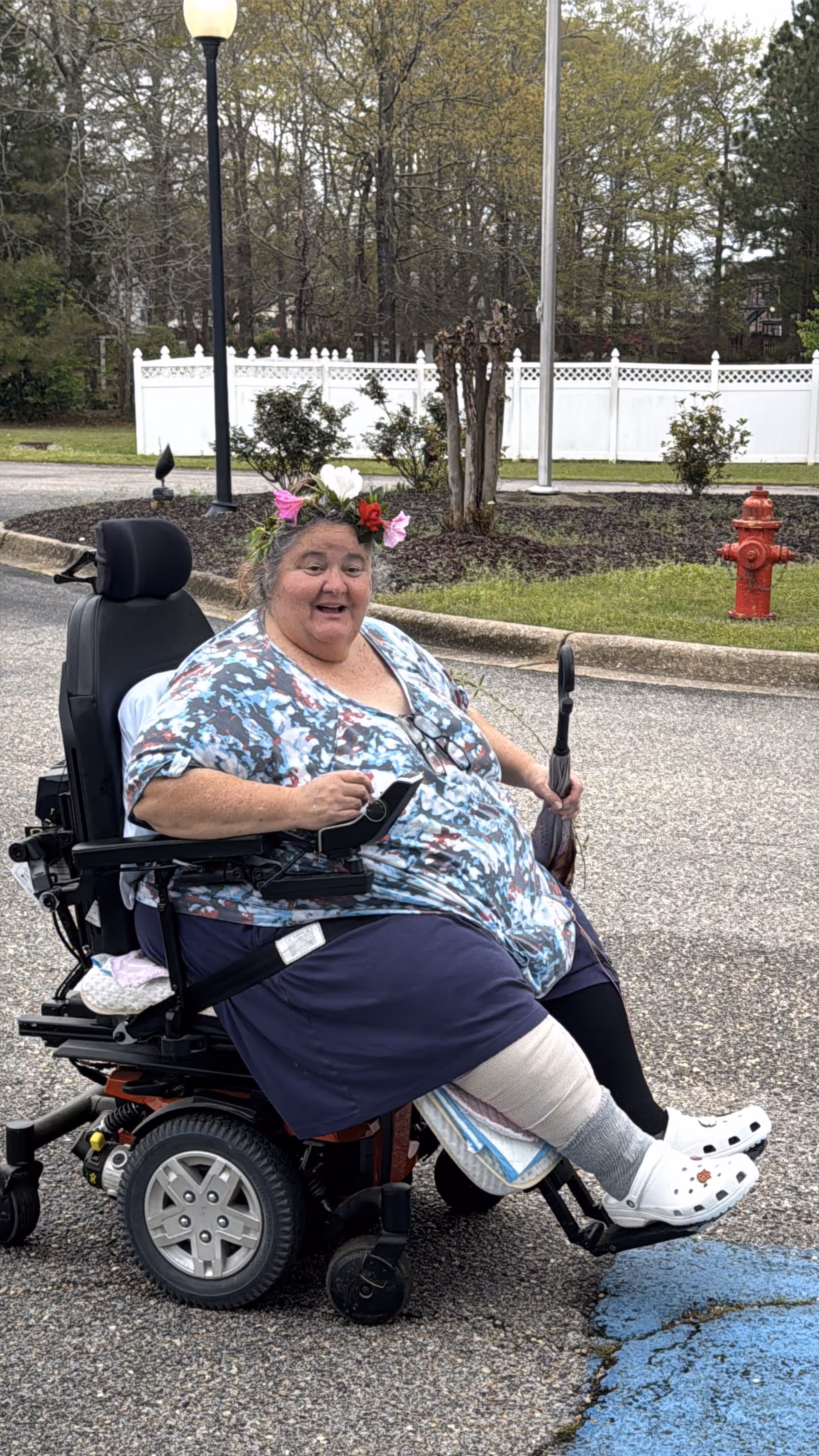 A smiling woman in a motorized wheelchair outdoors on a paved area near a garden with a white fence and trees in the background. She is wearing a floral headband, a patterned shirt, a dark skirt, and white Crocs. She has a bandage on her leg and is holding an umbrella.