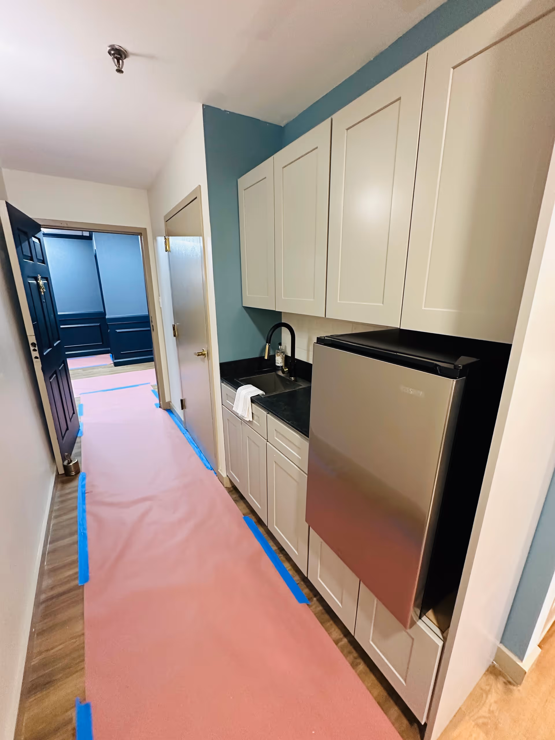 A narrow kitchenette area with white cabinets, a black countertop, a small stainless steel sink with a black faucet, and a compact stainless steel refrigerator. The floor is covered with a protective pink sheet taped down with blue tape. The walls are painted white and teal, and there are two doors visible at the end of the hallway.