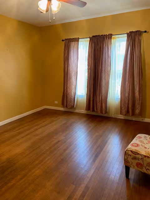 A spacious room with hardwood flooring, yellow walls, and large windows covered by brown curtains.