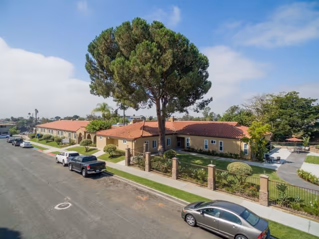 Exterior view of Arroyo Vista Nursing Center showing a single-story building with a red tile roof, beige walls, and a large tree in the front yard. Several cars are parked along the street in front of the facility. The area is landscaped with bushes, grass, and a fenced garden area. The sky is partly cloudy with blue patches.