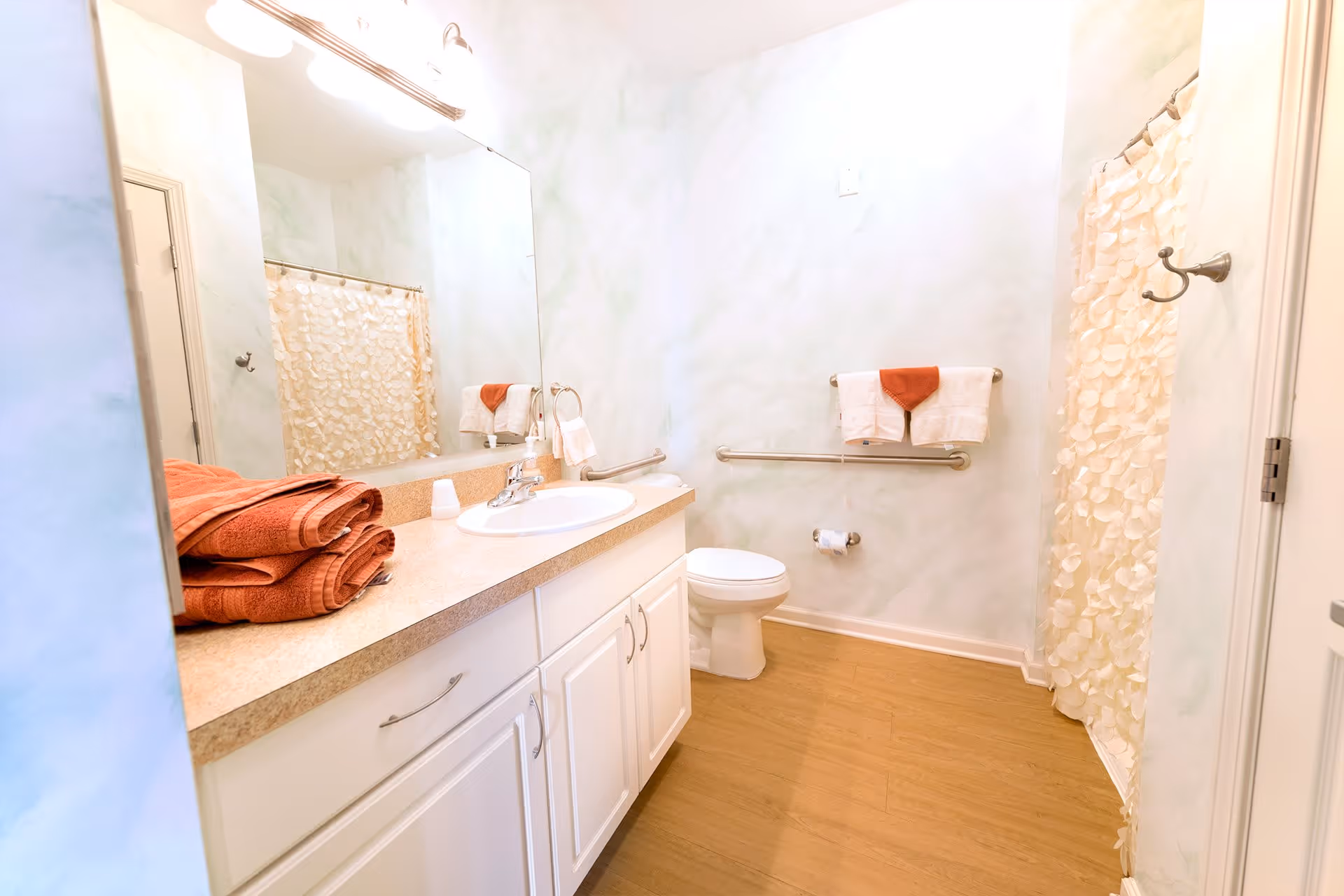 A clean bathroom with a white vanity and sink, a large mirror above the sink, a toilet with grab bars on the wall, a towel rack with white and orange towels, and a shower with a textured cream-colored curtain. The floor is wood-style, and the walls have a light, marbled pattern.