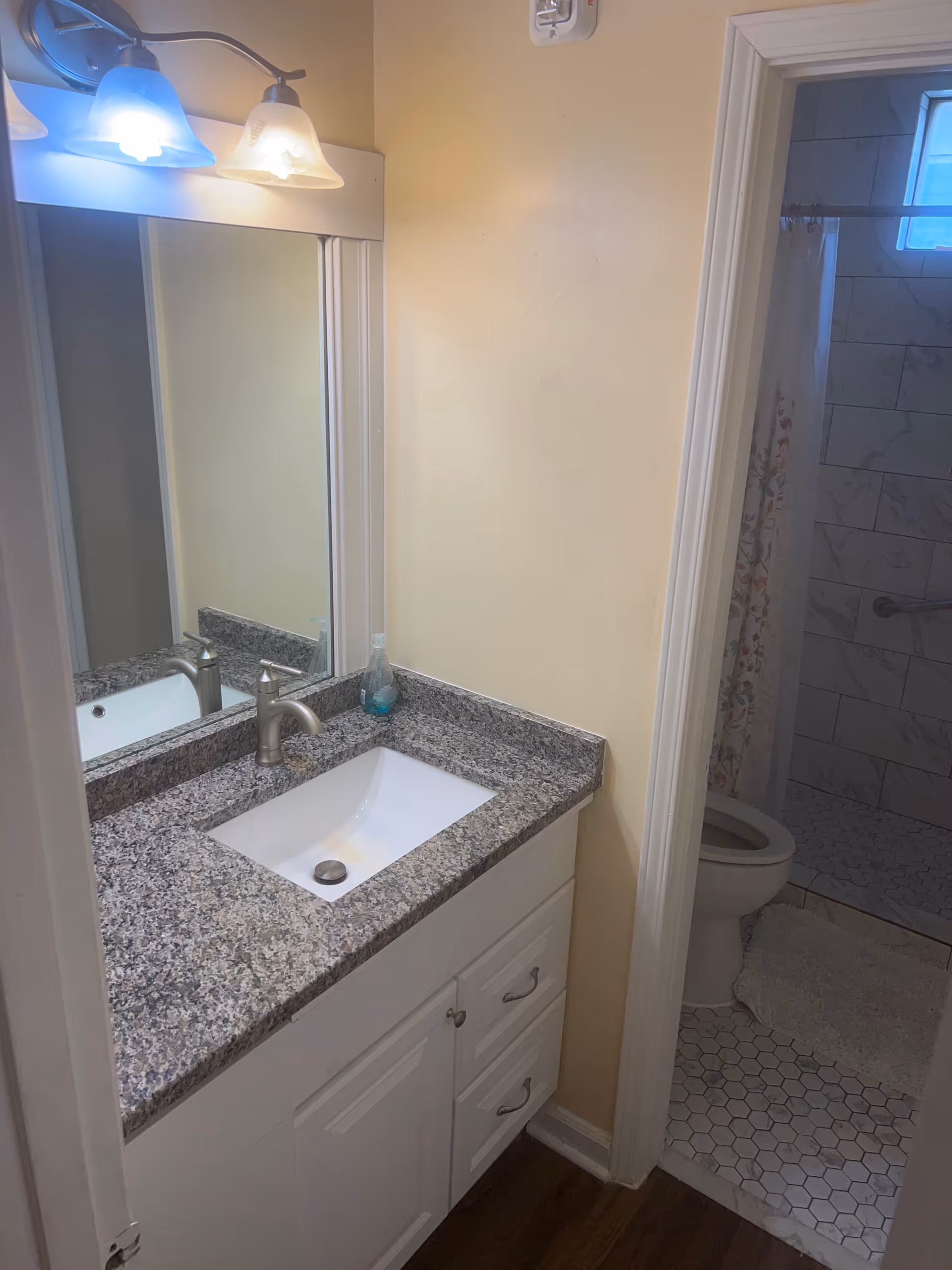A bathroom vanity with a granite countertop, a rectangular white sink, and a silver faucet. Above the sink is a large mirror with a three-light fixture. To the right, there is a doorway leading to a small bathroom area with a toilet, a shower with a floral curtain, and hexagonal floor tiles.
