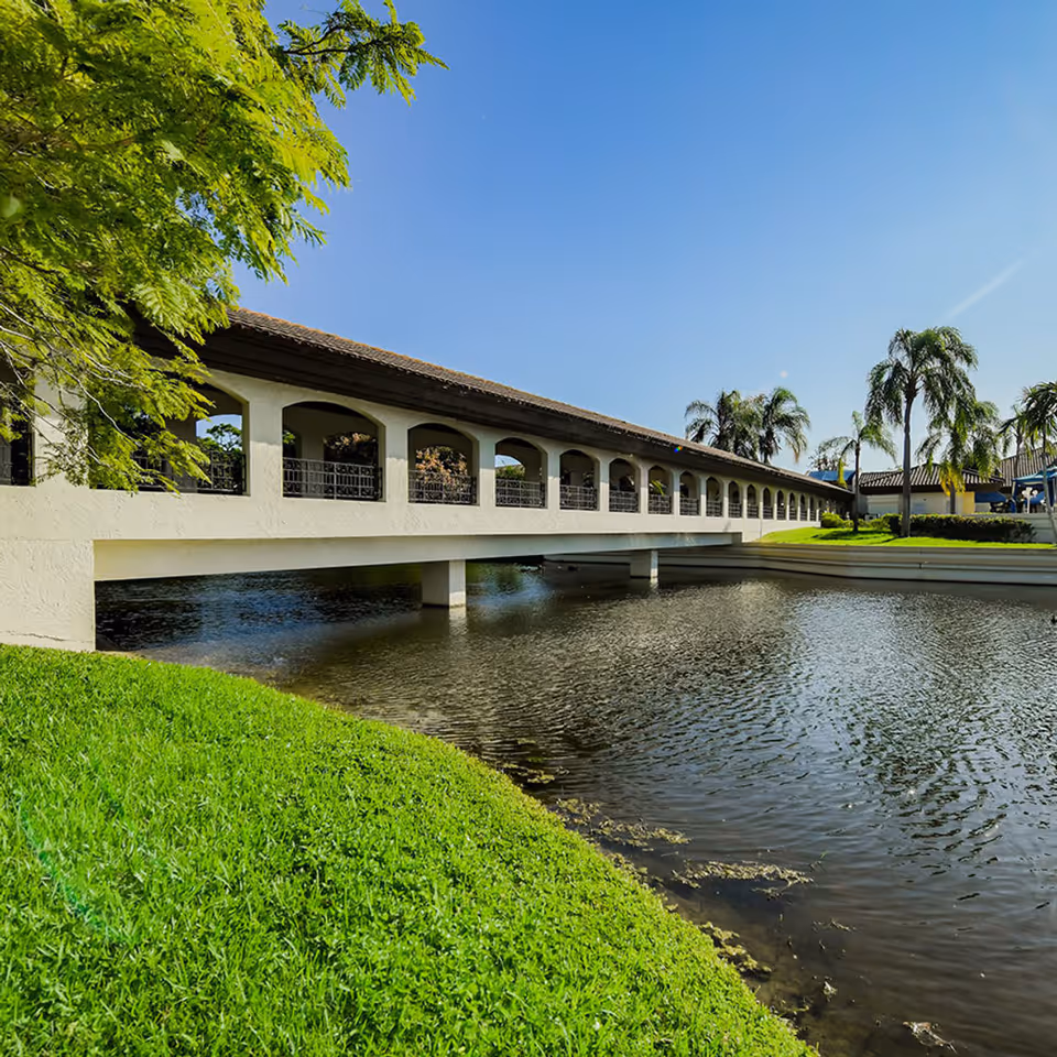 A covered walkway bridge with arches over a body of water, surrounded by green grass and palm trees under a clear blue sky.