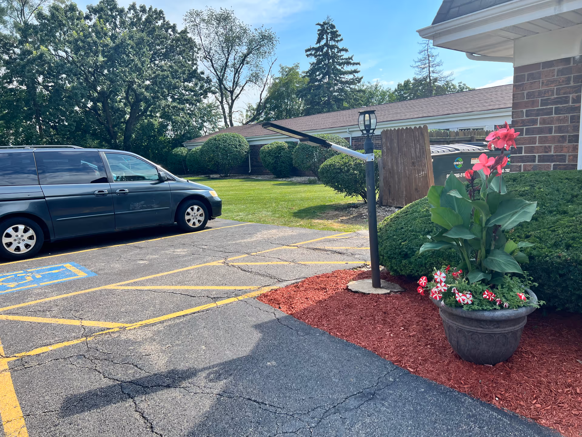 Parking area outside a building with a black minivan parked in a handicapped spot. There is a flower pot with pink and red flowers on a mulch bed next to a brick wall. Green bushes and trees are visible in the background under a blue sky.