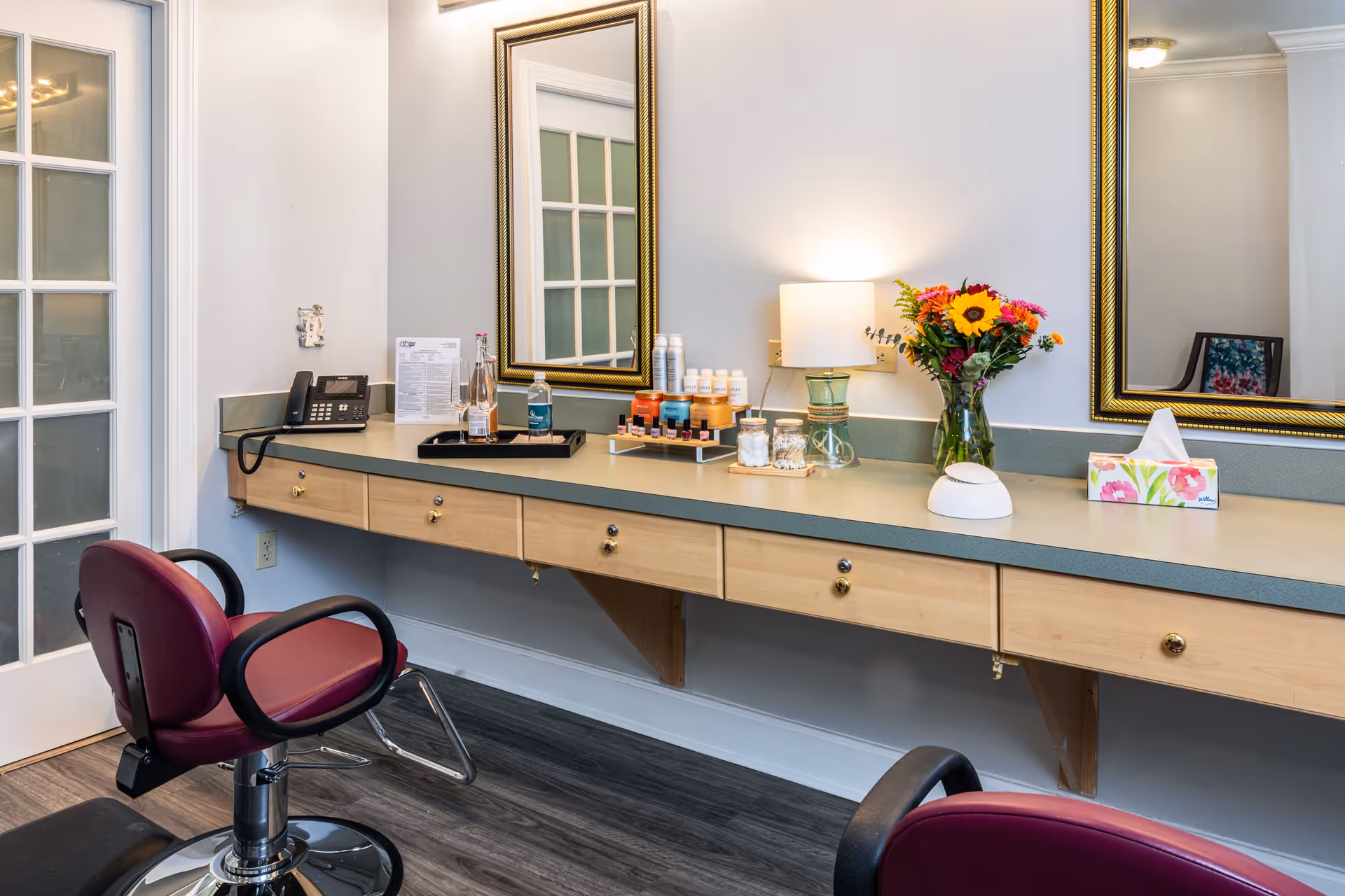 Interior view of a salon or grooming area with two maroon salon chairs facing a long counter with drawers. The counter holds various items including a telephone, bottles, a lamp, a vase with colorful flowers, and a tissue box. Two large mirrors with gold frames are mounted on the wall above the counter.