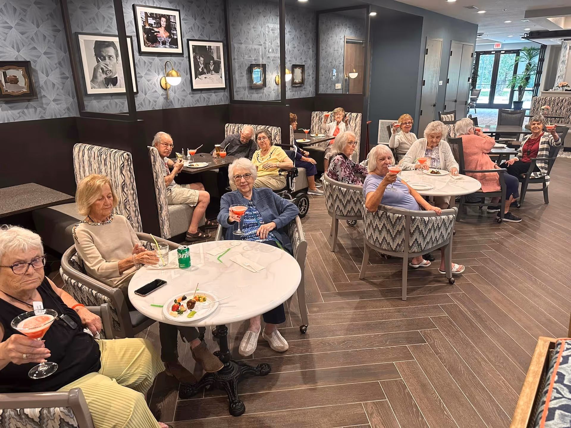 A group of elderly people sitting at tables in a communal dining or lounge area, holding up glasses with pink drinks in a toast. The room has patterned wallpaper, framed black and white photos on the walls, and large windows letting in natural light.