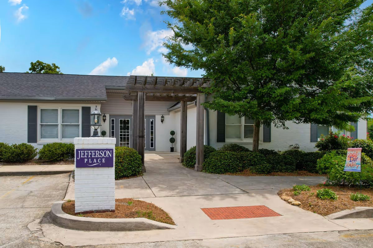 Front entrance of Jefferson Place with a white brick sign, covered pergola walkway, and surrounding shrubs and a tree.
