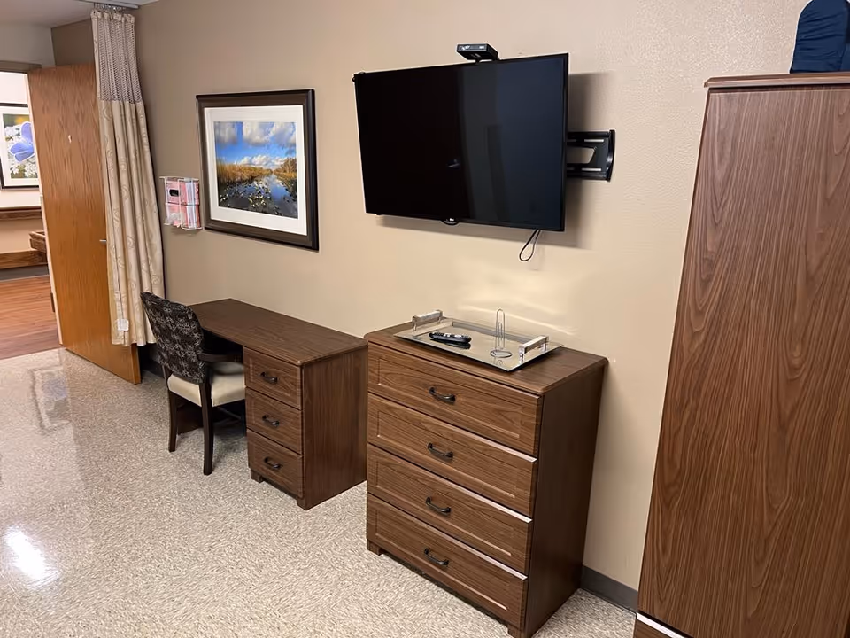 Interior view of a room with a wooden desk and chair, a framed landscape photograph on the wall, a flat-screen TV mounted above a wooden dresser with a tray and remote control on top, and a wooden wardrobe on the right side.