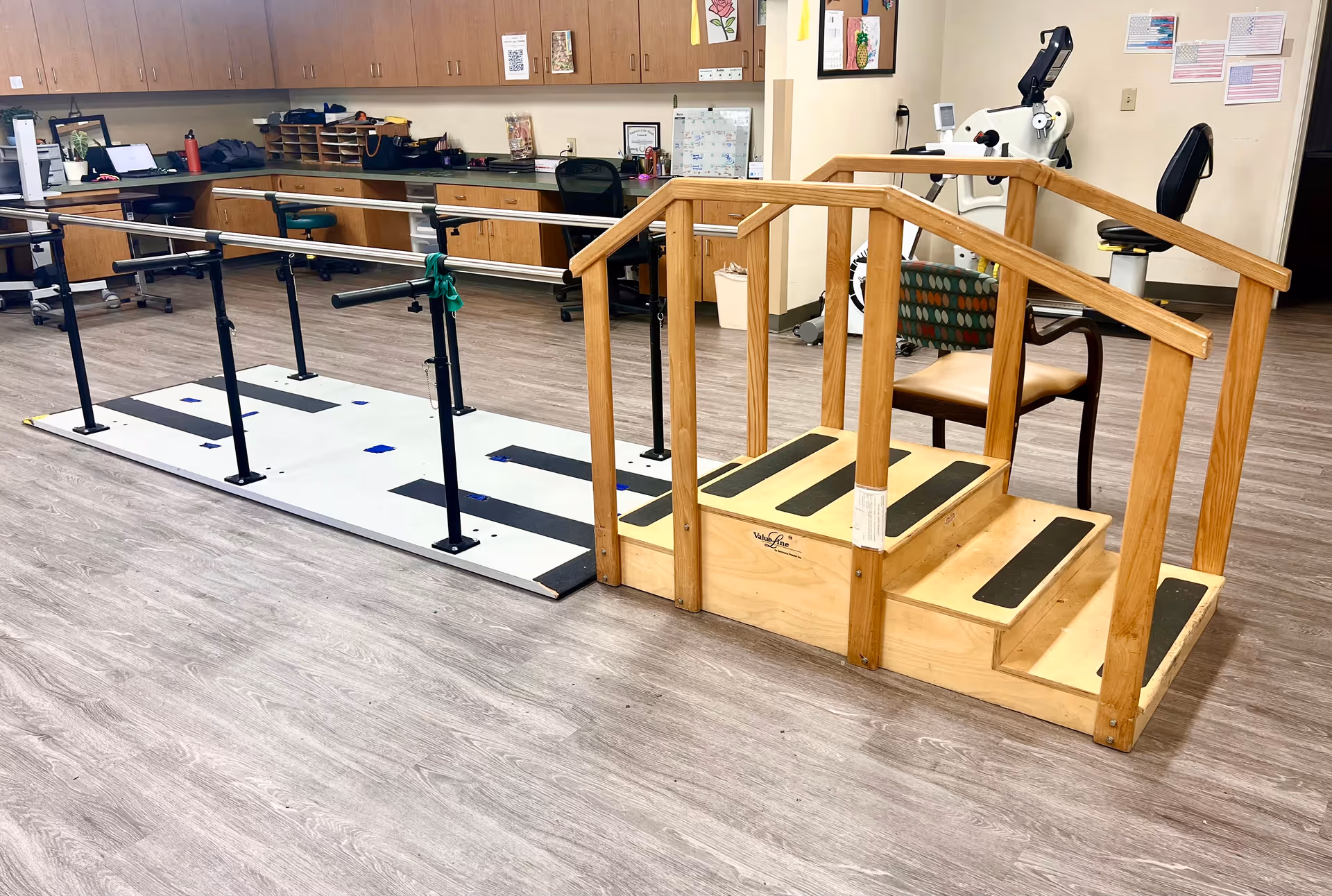 A rehabilitation therapy room with parallel bars for walking exercises and a wooden staircase with handrails for physical therapy. The room has wood flooring, cabinets along the back wall, a desk with a chair, and exercise equipment in the background.