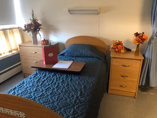 A single bed with a blue quilted bedspread in a room with two wooden nightstands on either side. Each nightstand has decorative autumn-themed flower arrangements. There is a window with curtains on the left side and a wall-mounted light fixture above the bed. A small table tray with a red gift bag and a notebook is placed on the bed.