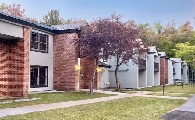 Exterior view of a senior living facility building with brick and light gray walls, multiple windows, and small trees along a paved walkway surrounded by grass and greenery.