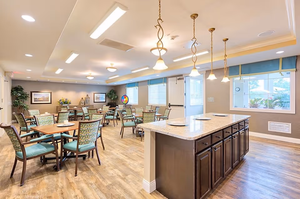 A bright and spacious dining room with multiple wooden tables and chairs arranged neatly. The room features a large island with a granite countertop in the foreground, pendant lights hanging from the ceiling, large windows with blue valances allowing natural light, and framed artwork on the walls. The floor is wooden, and there are some plants and decorative items in the background.