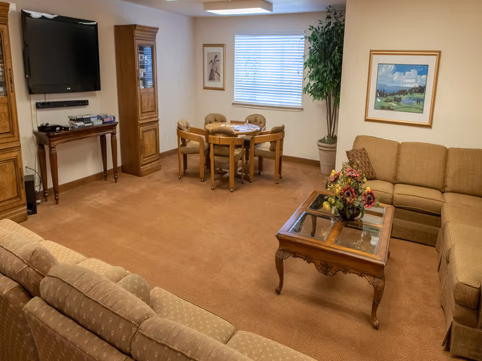 A cozy living room in a senior living community featuring a large beige sectional sofa, a wooden coffee table with a floral arrangement, a round table with four cushioned chairs near a window with blinds, a flat-screen TV mounted on the wall, and wooden cabinets. The room is carpeted and decorated with framed artwork and a potted plant.