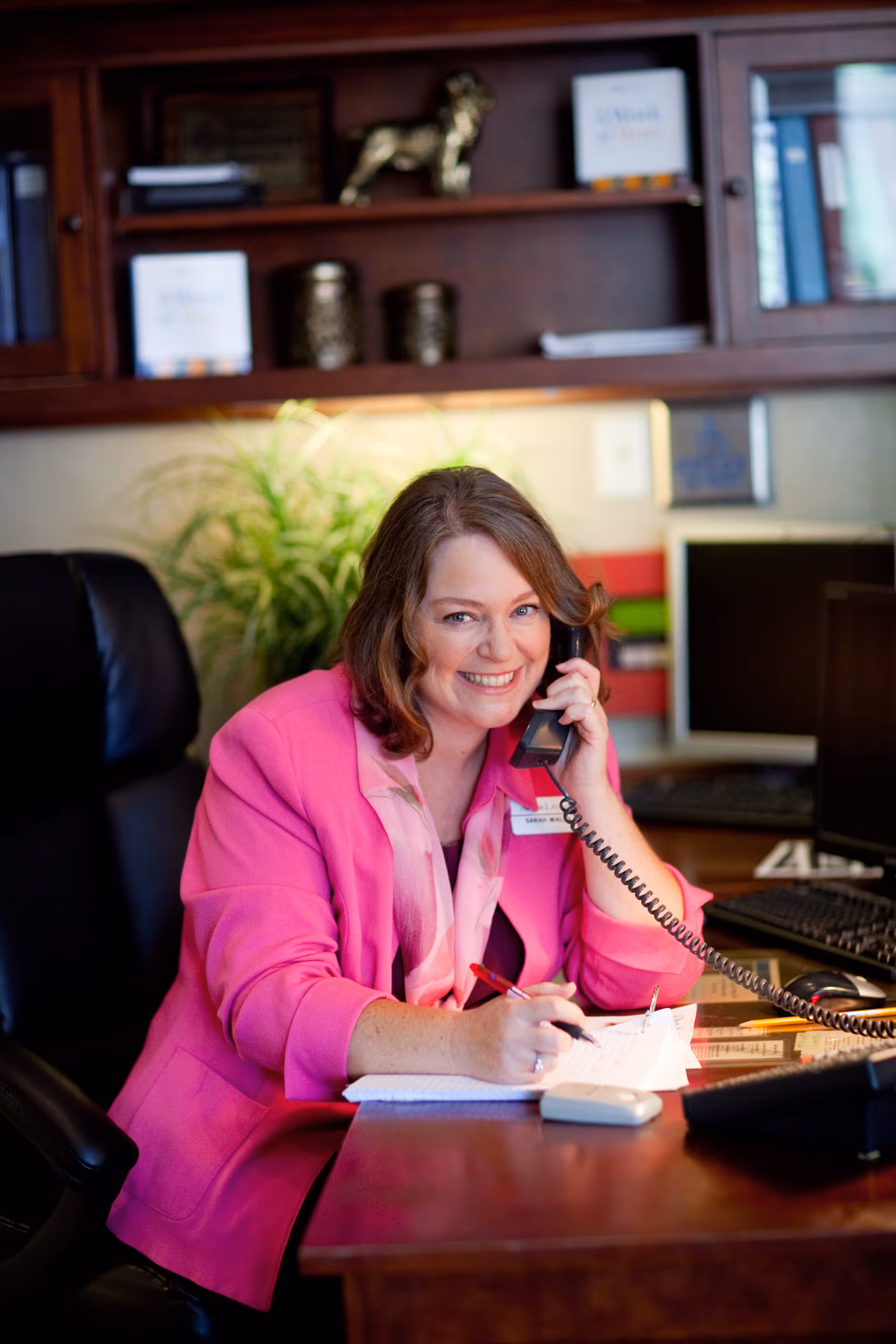 A woman wearing a bright pink blazer is sitting at a desk in an office, smiling while talking on a corded phone and writing on a notepad. Behind her are wooden shelves with books and decorative items, and a green plant is visible in the background.