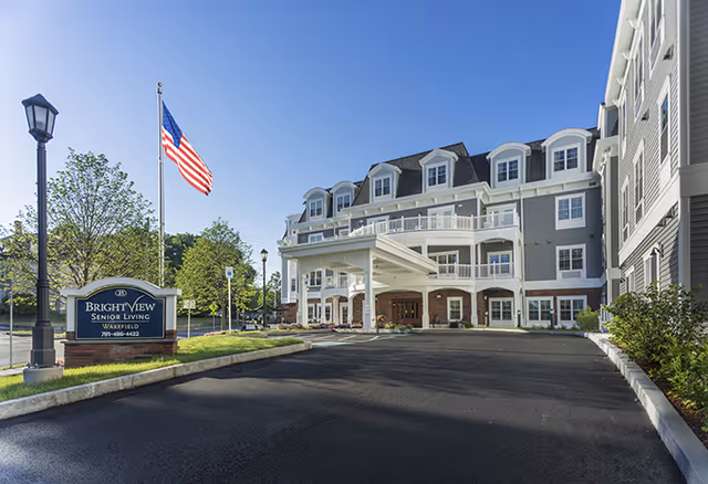 Exterior view of Brightview Wakefield senior living facility with a large driveway, an American flag on a flagpole, and a sign displaying the facility name. The building is multi-story with balconies and a covered entrance.