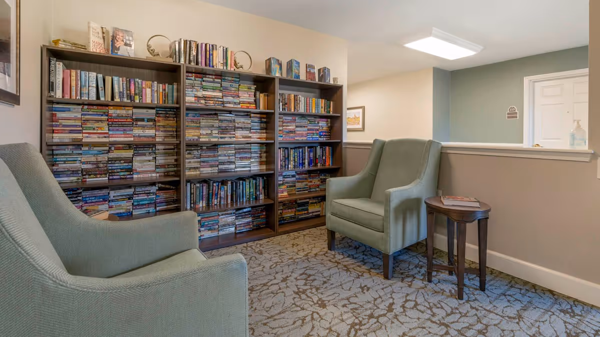 A cozy reading nook in a senior living facility featuring two light green upholstered armchairs facing each other with a small wooden side table between them. Behind the chairs are three tall bookshelves filled with numerous books. The room has neutral-colored walls and a patterned carpet.