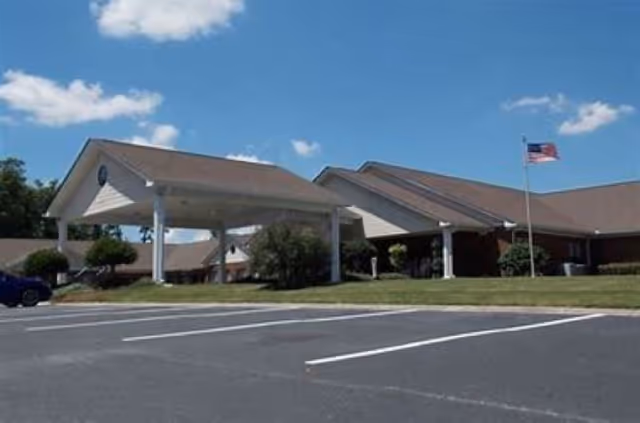 Exterior view of a single-story senior living facility building with a covered entrance, an American flag on a flagpole, and a parking lot in the foreground under a blue sky with some clouds.