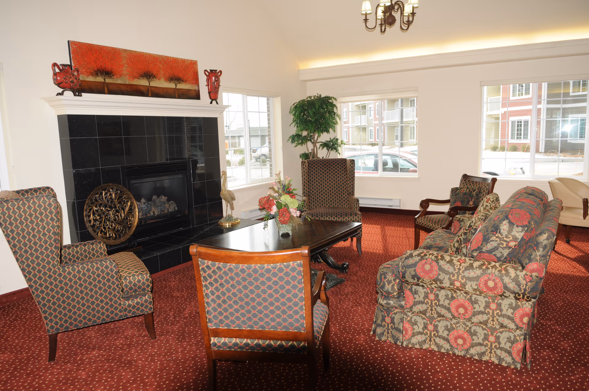 A cozy living room area with patterned upholstered chairs and a floral sofa arranged around a dark wooden coffee table. There is a black tiled fireplace with decorative items on the mantel, including a red painting and two red vases. Large windows let in natural light and show a view of parked cars and buildings outside. The room has a red carpet and a chandelier hanging from the ceiling.
