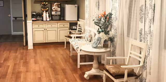 Cozy interior seating area with wood floors, cream-colored chairs and a round side table topped with flowers, and a small kitchenette with a coffee station in the background.