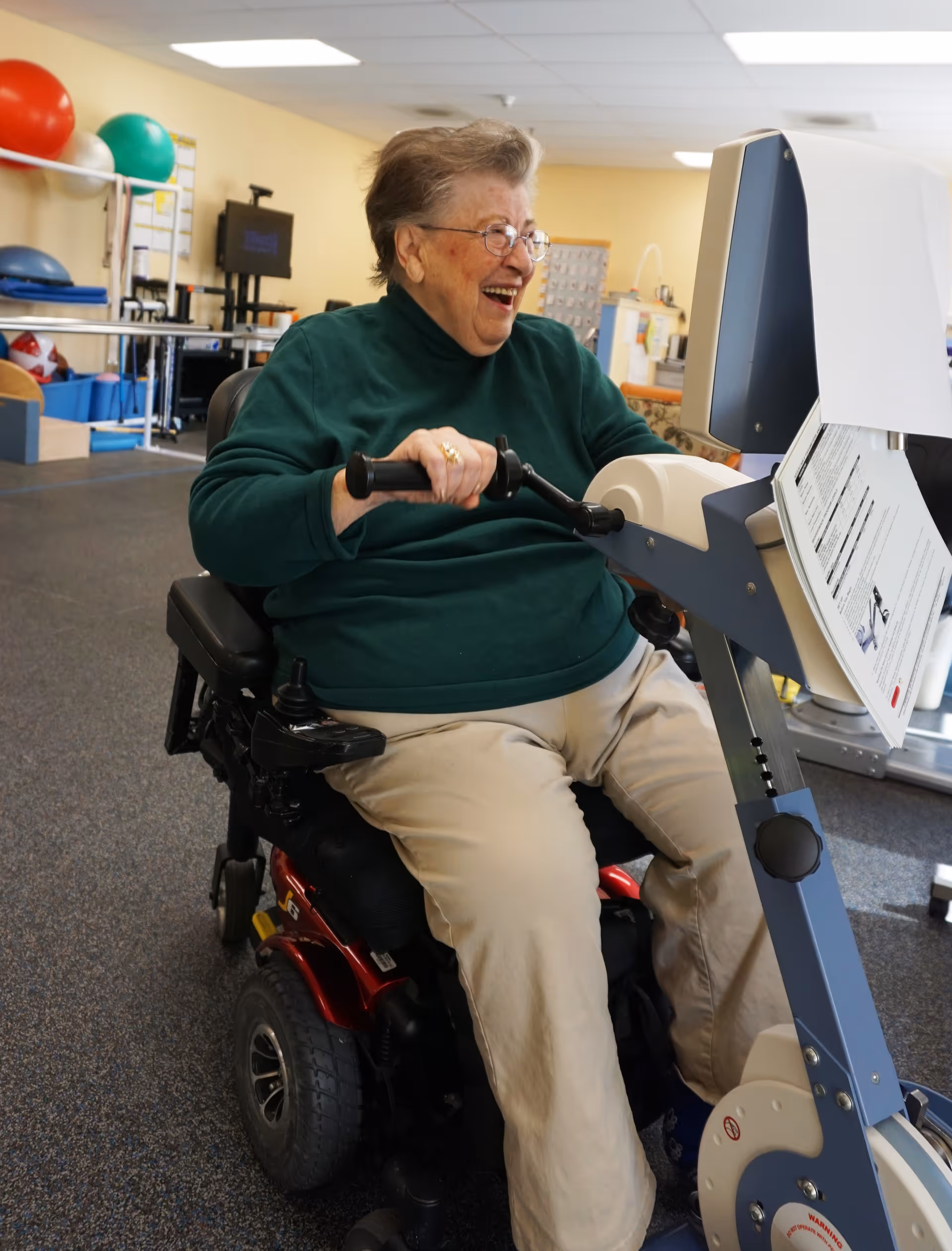 An elderly woman in a green sweater and beige pants is seated in a motorized wheelchair, smiling and using a piece of exercise equipment in a rehabilitation or therapy room with various physical therapy tools and exercise balls in the background.