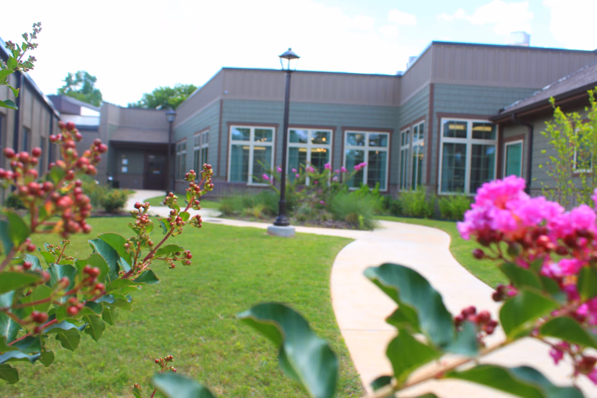 View of a senior living facility's outdoor garden area with a curved concrete pathway, green grass, flowering plants, and a building with large windows in the background under a partly cloudy sky.