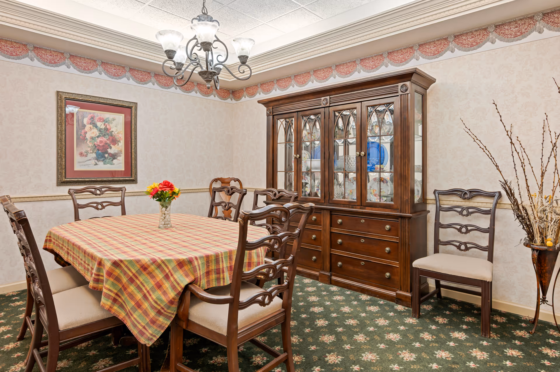 A traditional dining room with a plaid-covered table surrounded by wooden chairs, a glass-front china cabinet, chandelier, and floral decorations.