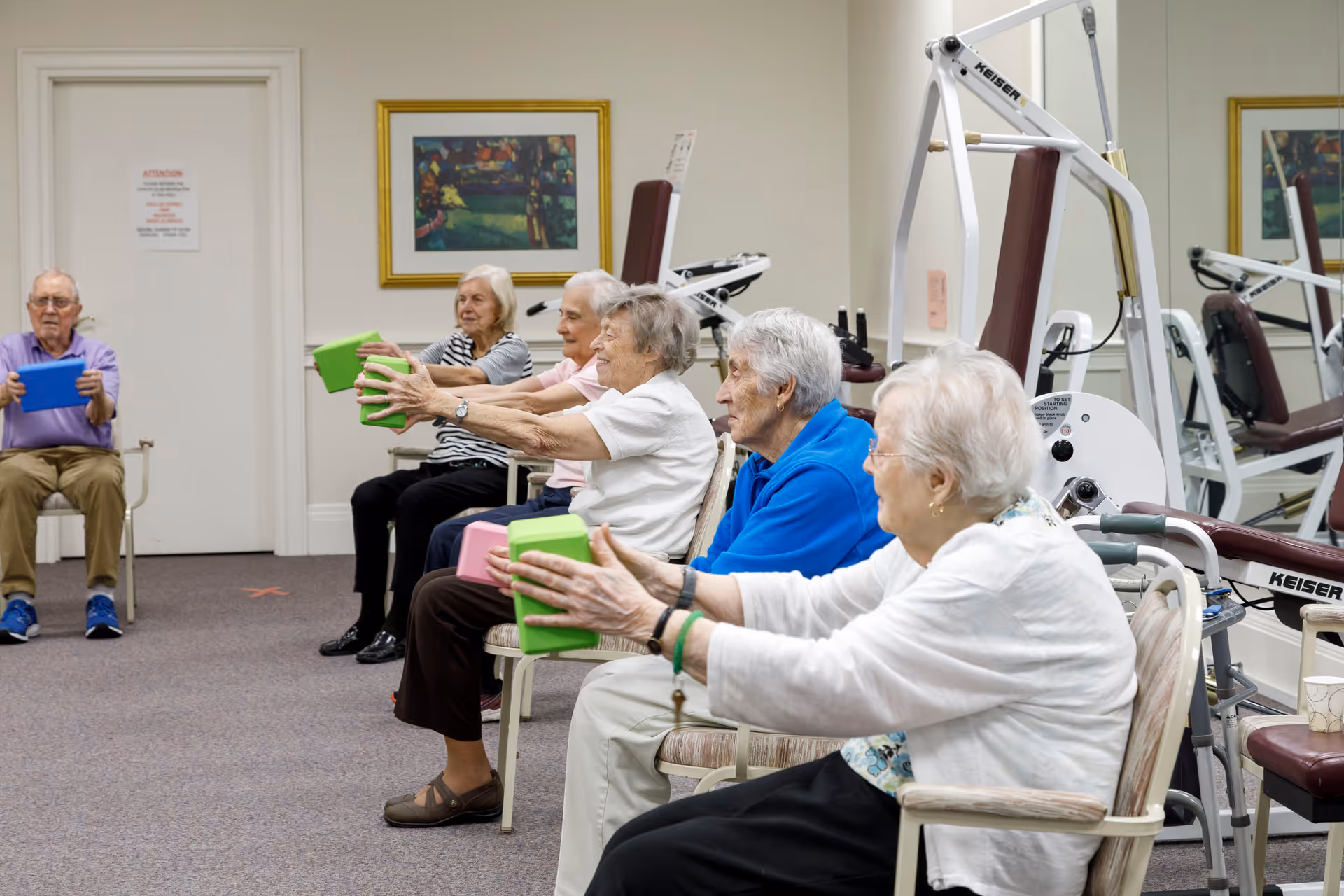 A group of elderly individuals seated in chairs in a fitness room, participating in a seated exercise class using colorful foam blocks. Exercise equipment and a large mirror are visible in the background.