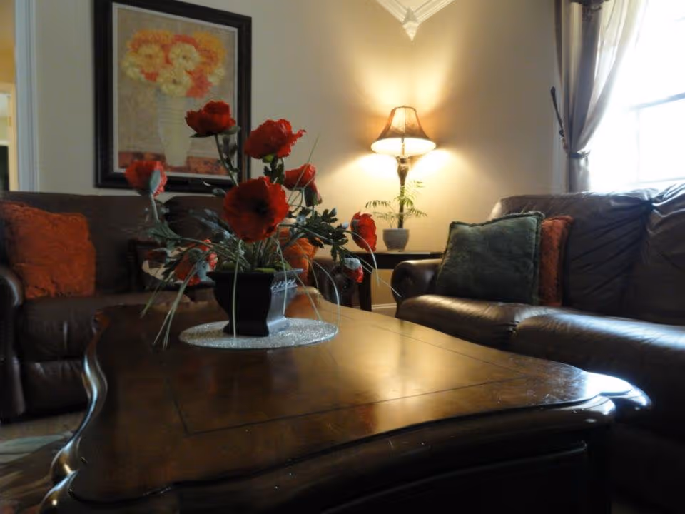 A cozy living room with brown leather sofas adorned with orange and green cushions, a wooden coffee table with a vase of red flowers, a side table with a lamp, and a framed painting of flowers on the wall.