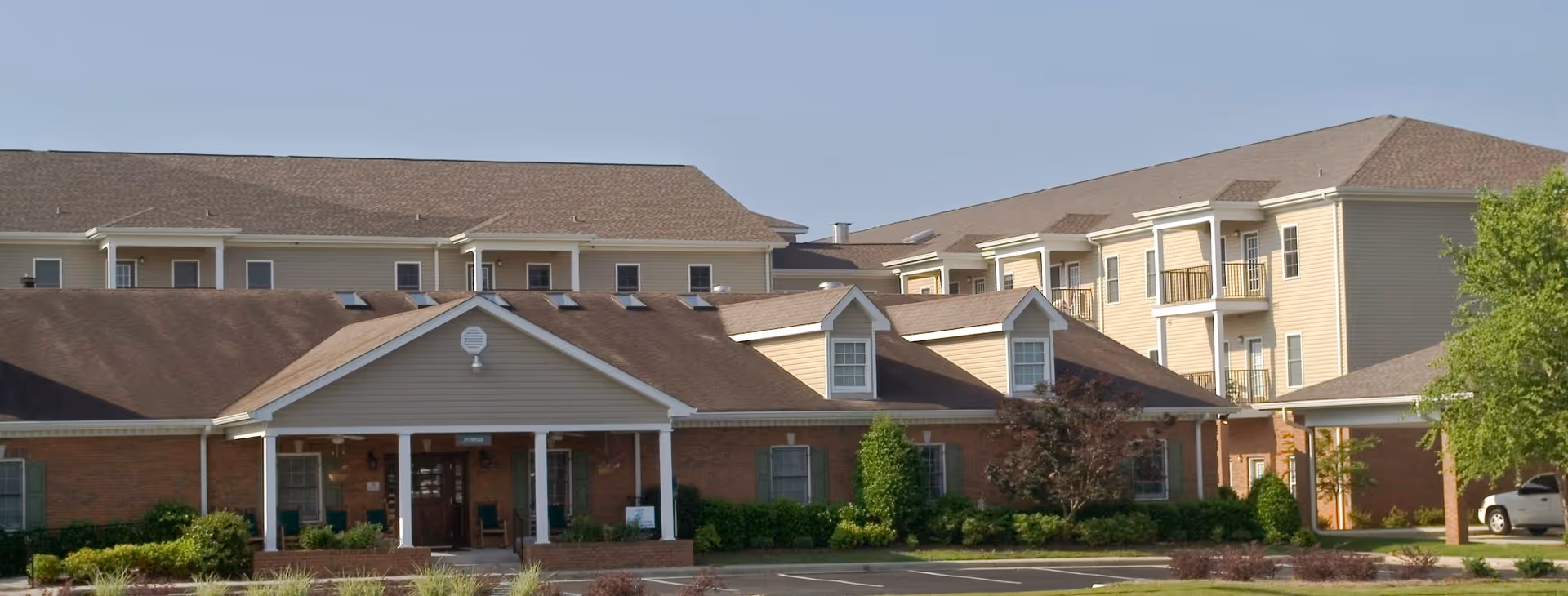 Exterior view of a multi-story senior living facility with beige siding and brick accents, featuring a covered entrance, balconies, and surrounding greenery under a clear sky.