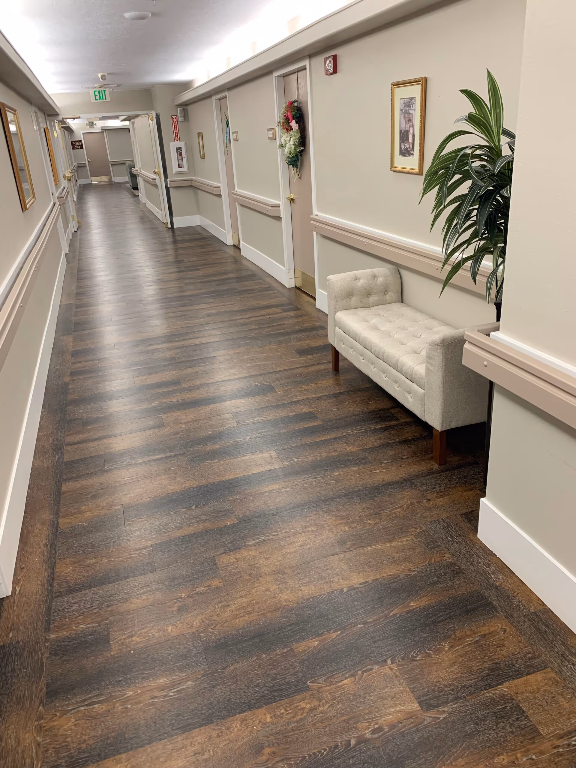 A long, clean hallway in an assisted living facility with wood-patterned flooring, beige walls, and several closed doors. There is a small cushioned bench and a potted plant along the right wall, with framed pictures hanging above. Exit signs and safety equipment are visible at the far end of the hallway.