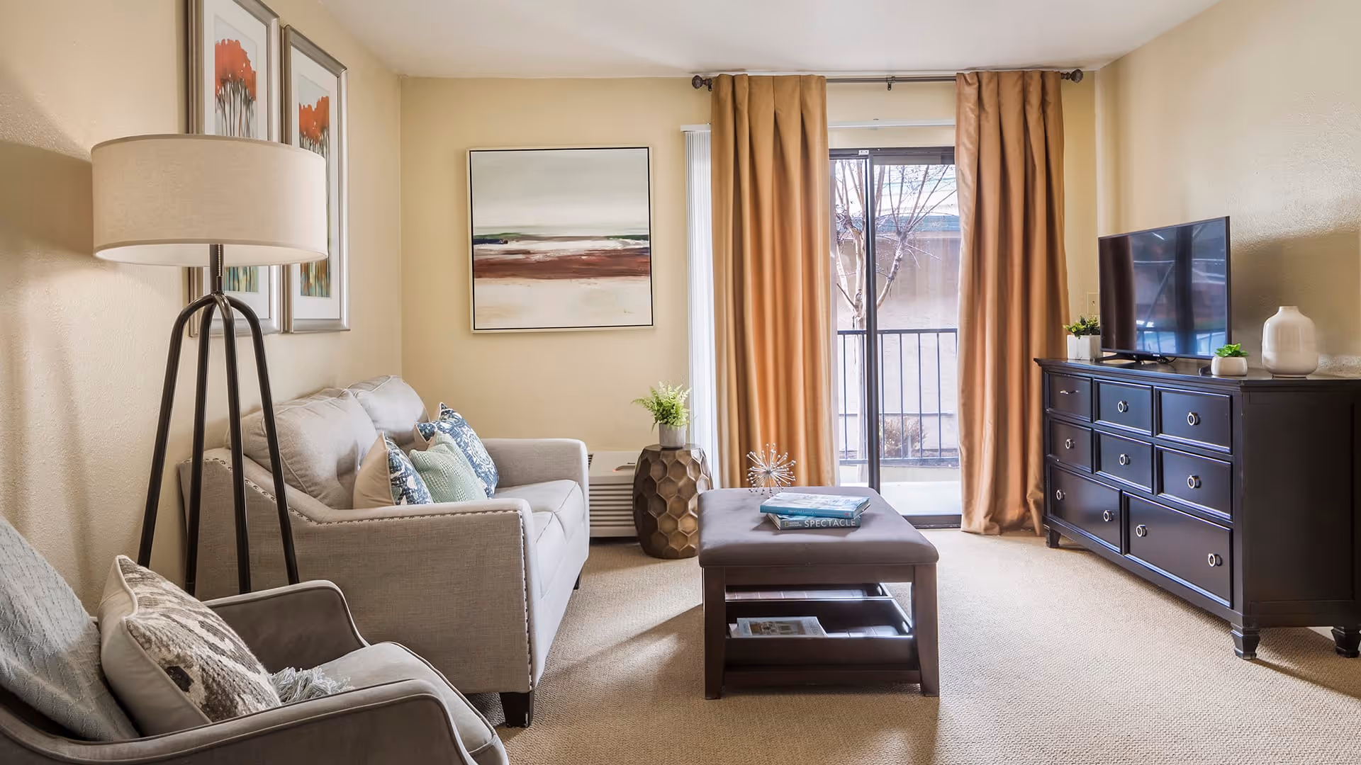 A cozy living room with a beige sofa adorned with patterned cushions, a gray armchair with a pillow, a tall floor lamp, and a dark wooden coffee table with books on it. There is a large window with brown curtains letting in natural light, and a dark wooden dresser with a flat-screen TV and decorative items on top. The walls are light-colored with framed artwork.