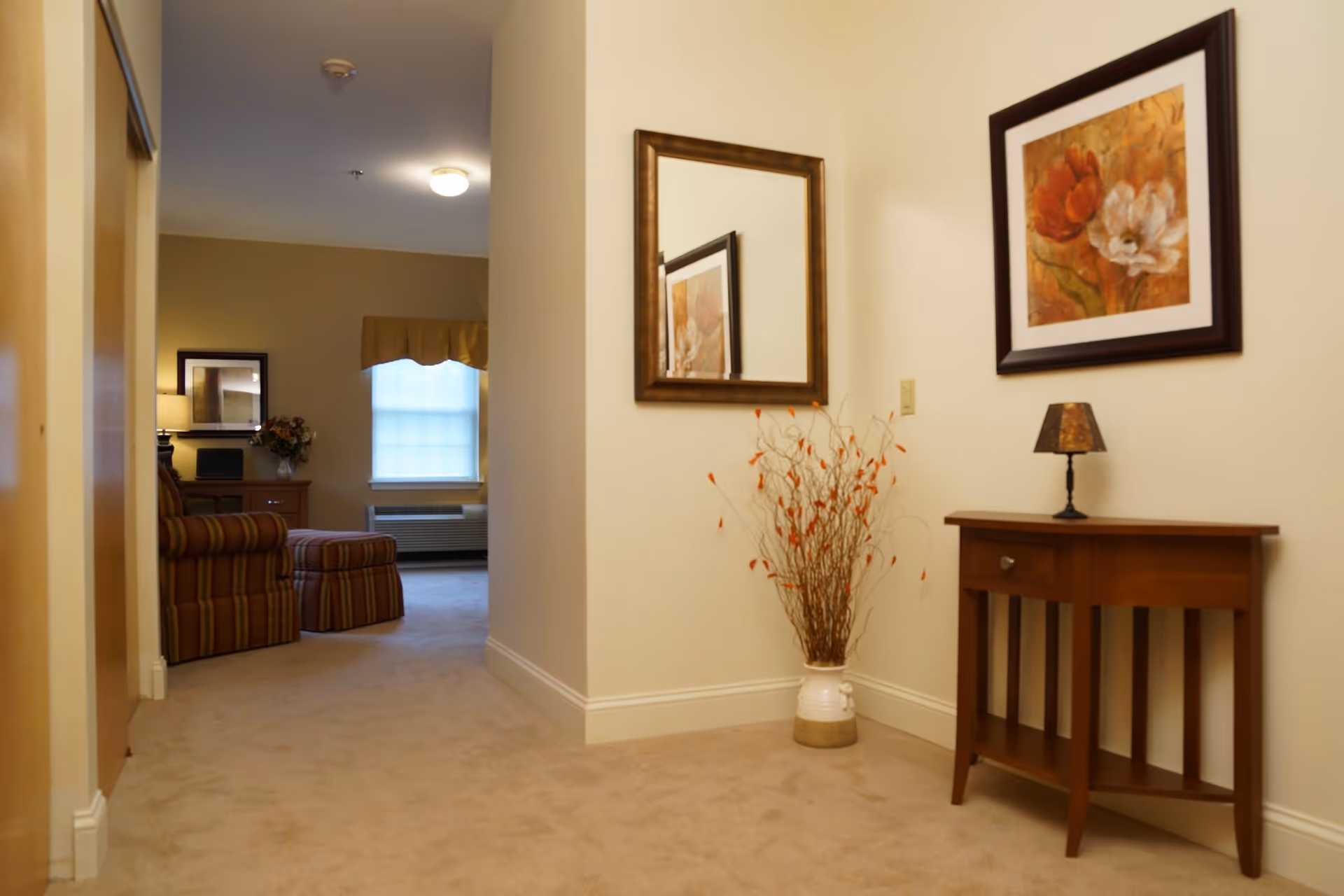 View down a carpeted hallway leading to a living room area with a striped armchair and ottoman. The hallway has a wooden console table with a small lamp and a vase with decorative branches. A framed floral painting and a mirror hang on the walls.
