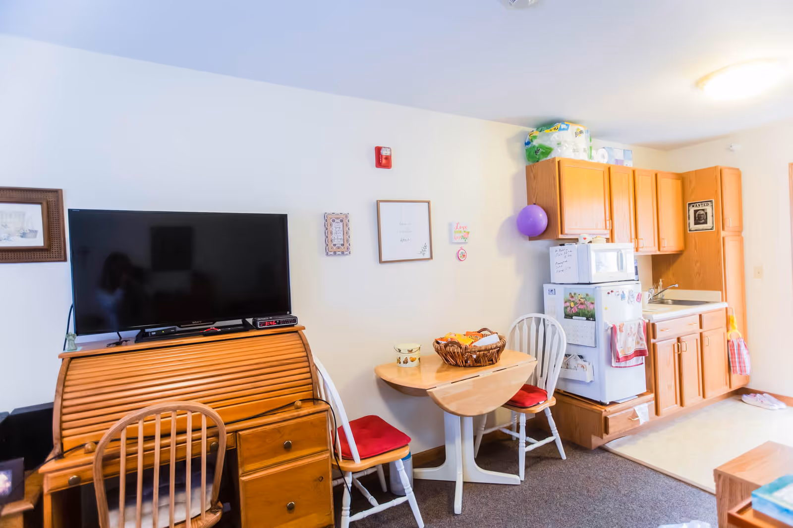A small living area and kitchenette in an assisted living facility. The room features a wooden roll-top desk with a flat-screen TV on top, two wooden chairs with red cushions, a small wooden drop-leaf table with a basket on it, and a kitchenette with wooden cabinets, a white refrigerator, microwave, and a sink. The walls are decorated with framed pictures and a purple balloon is attached to one of the cabinets.