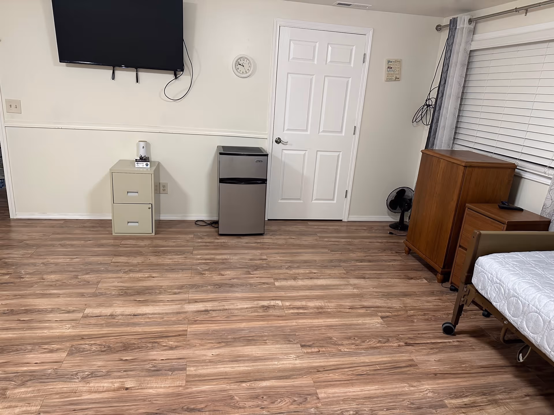 A room with wood flooring featuring a wall-mounted flat screen TV, a small beige filing cabinet with a tissue box on top, a compact stainless steel mini fridge, a white door, a wall clock, a wooden dresser and nightstand, a small black fan, and a bed with a white quilted bedspread partially visible on the right side. The room has white walls and a window with closed blinds and gray curtains.